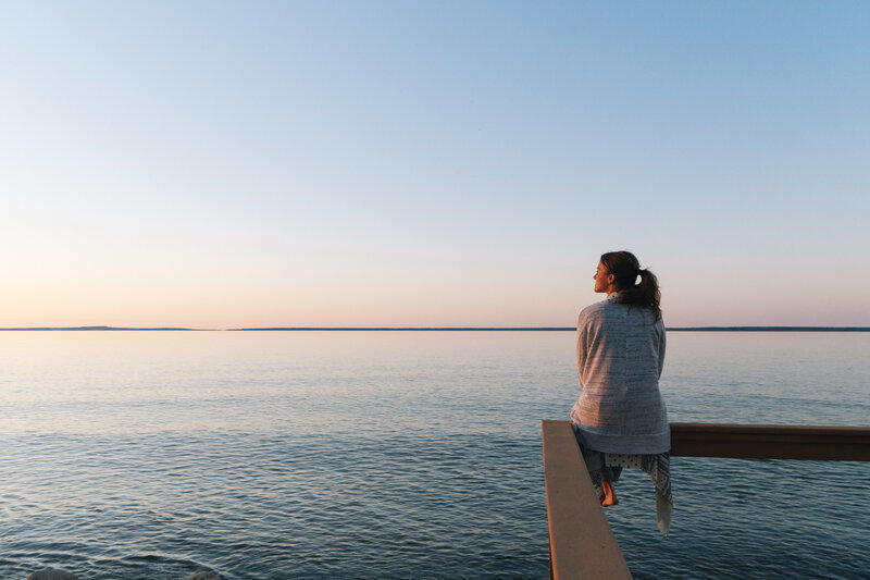 A young woman gazes off towards the sunset while she sits on an edge overlooking water/