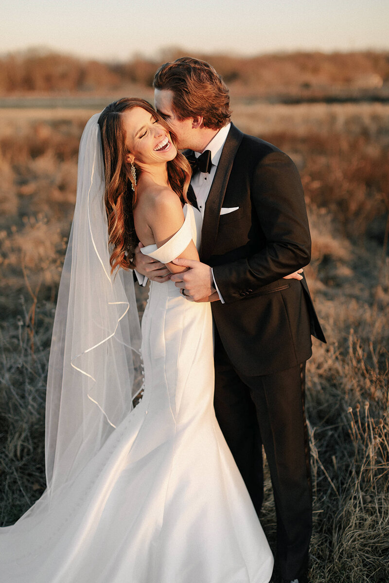 groom embracing bride in wedding dress and veil as she laughs