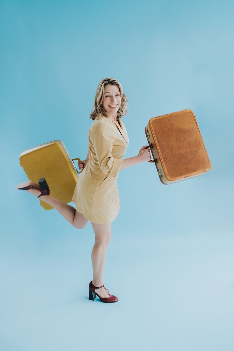 Woman with shoulder-length blonde hair holds two vintage suitcases while smiling and posing like she is running.