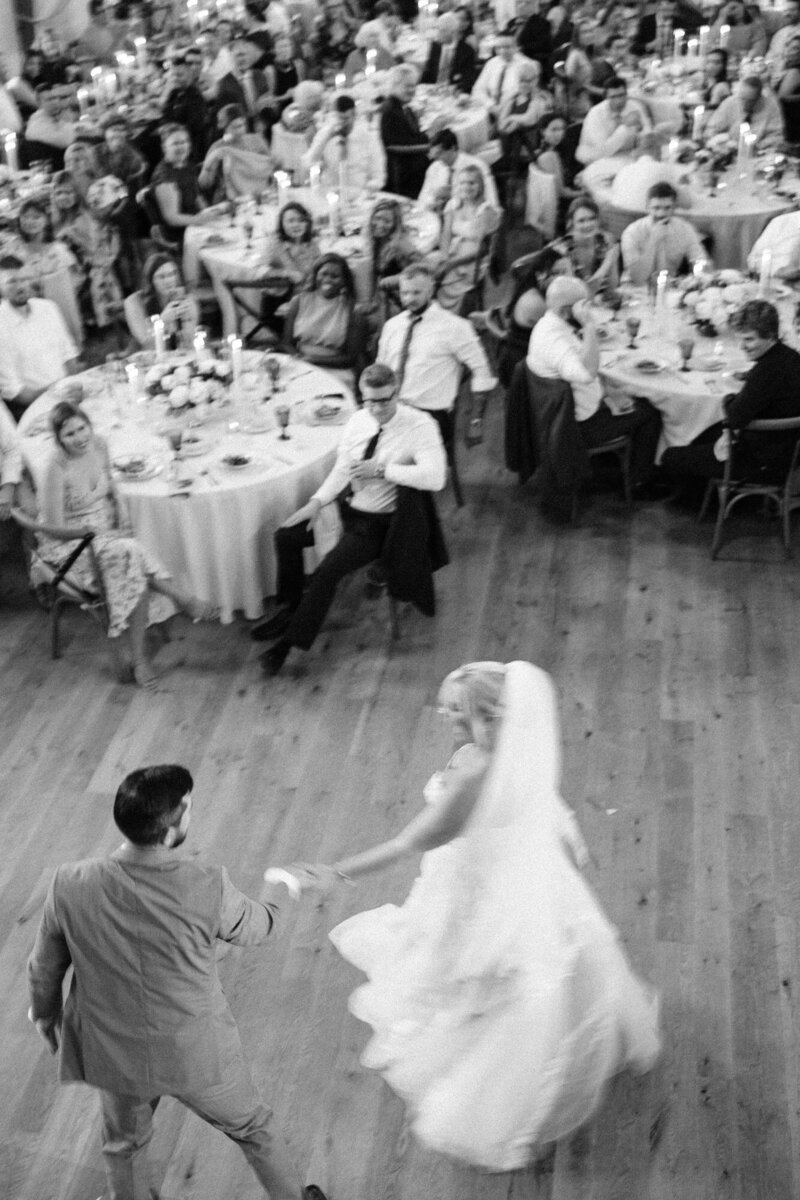 A bride and groom dancing in the foreground with guests seated at tables in the background, in a black and white photo capturing the motion of the moment.