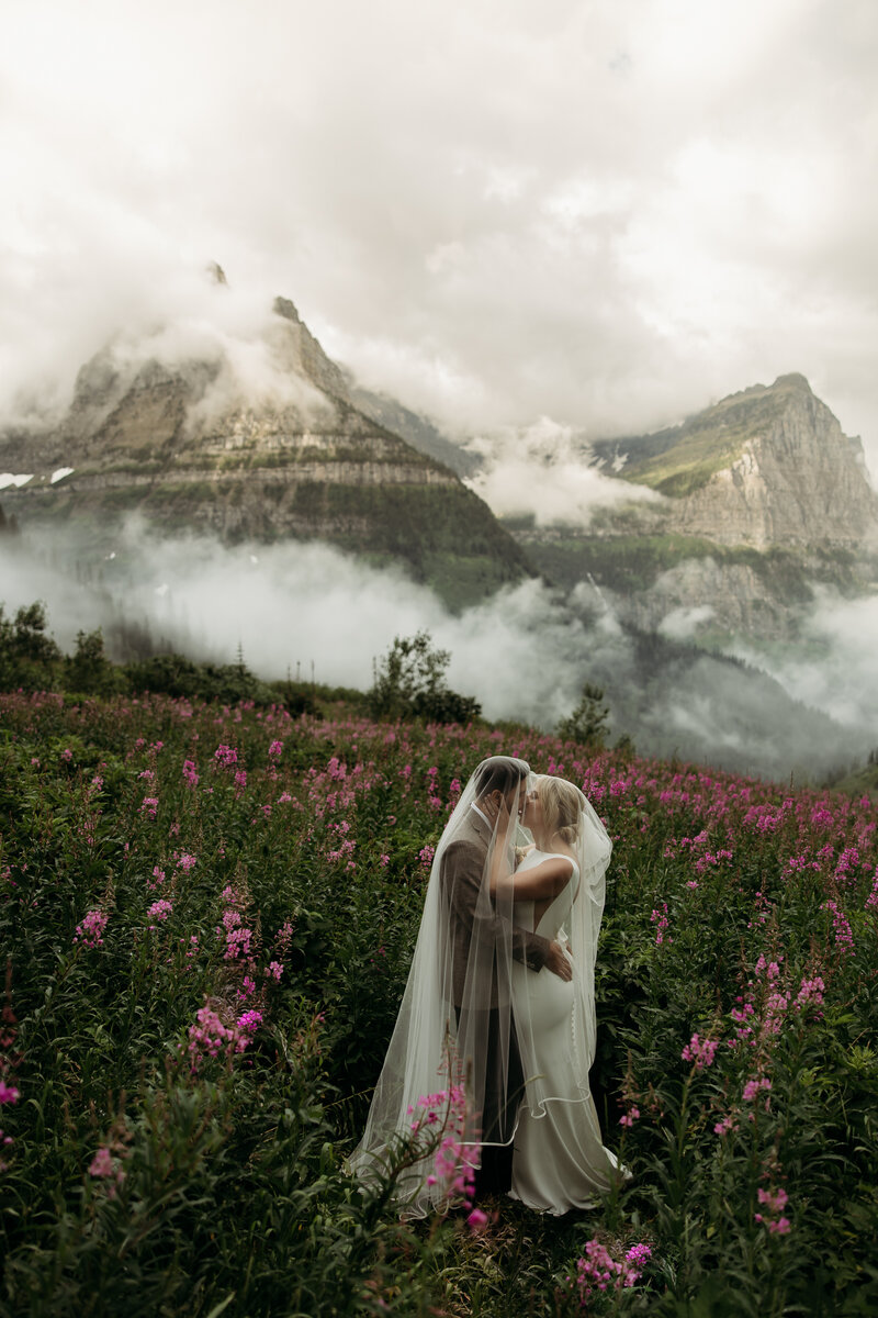 a bride and groom sitting in wildflowers surrounded by glacier national park mountains