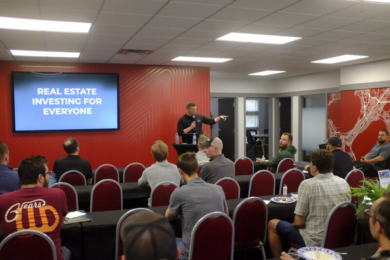 Person giving presentation to a room of people.  Screen on wall says "Real Estate Investing for Everyone"