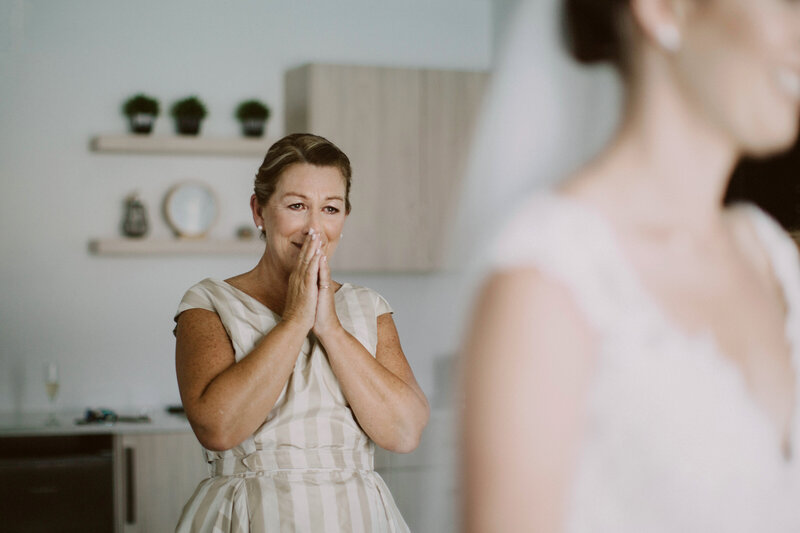Mother of the bride caught in a candid moment of real emotion as she sees the bride. Her hands over her mouth and she has tears in her eyes.