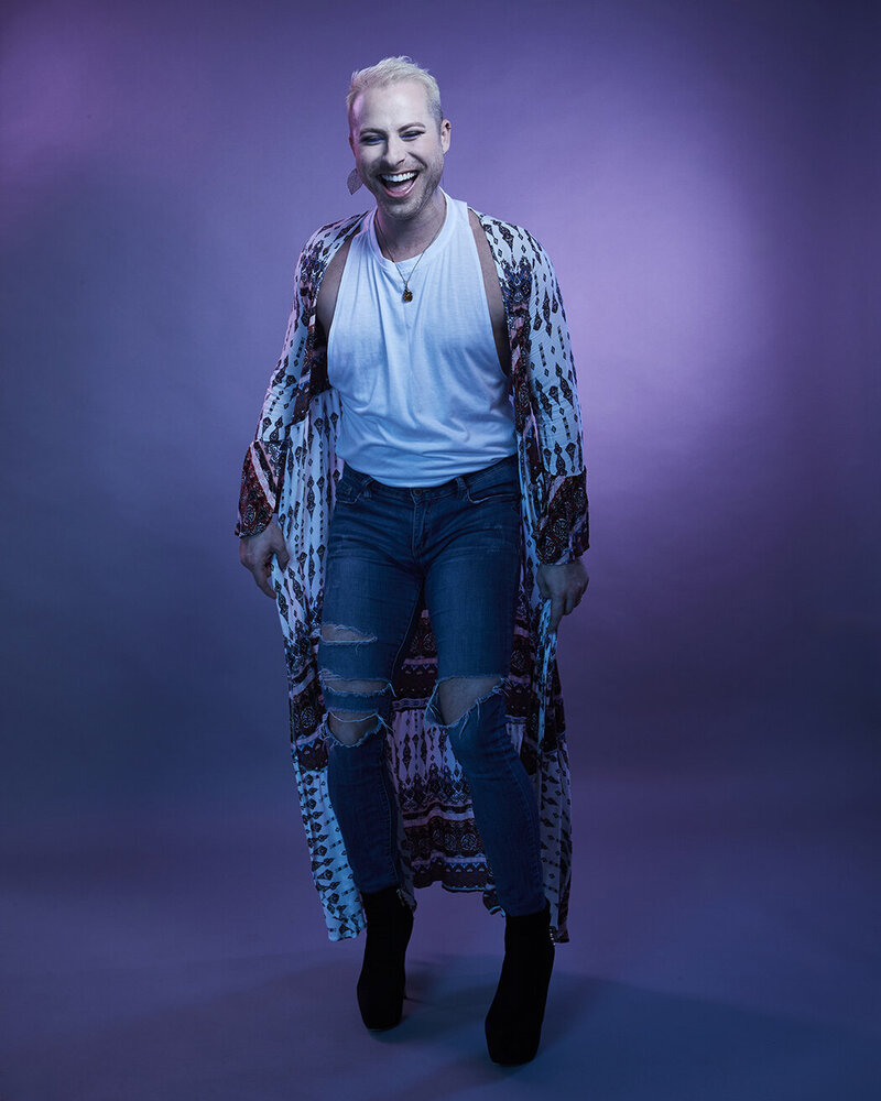 Full-body portrait of Dain August wearing a white outfit with black patterns, striking a bold pose in a studio setting, captured by Ylva Erevall.