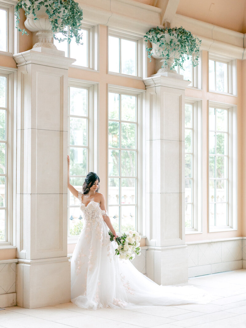 Bride in a white wedding dress with pink appliques on the bodice and skirt looking over her shoulder leaning against a pillar with greenery in urns on top of the pillars
