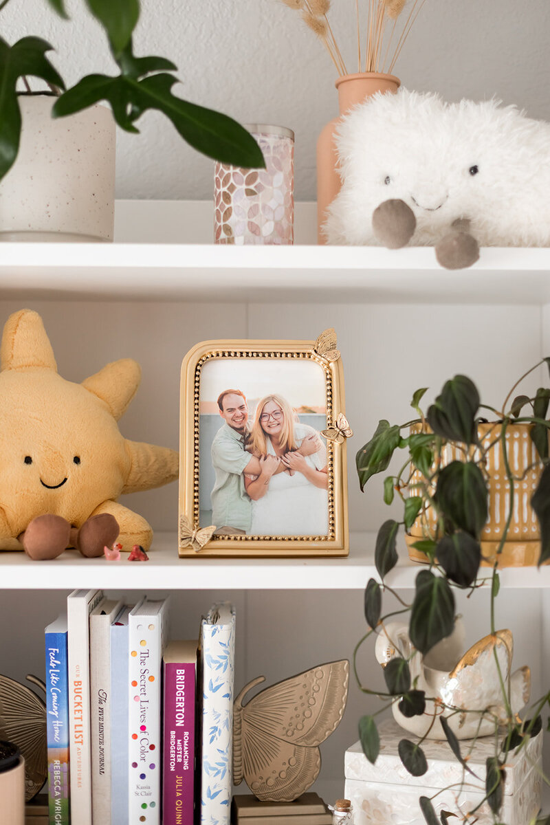 Gold frame on white shelf showing couple embracing surrounded by house plants, books, and jellycat decor