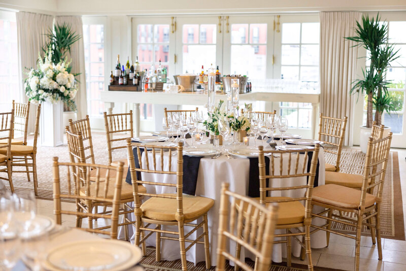 A round dining table set for a formal event with white linens, elegant glassware, and floral centerpieces. Surrounded by wooden chairs, the room features large windows, soft curtains, and a background with a bar area displaying bottles and glasses.