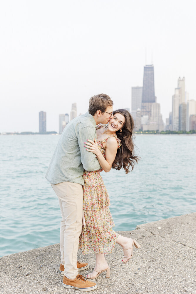 man kissing woman on the cheek by the water with a cityscape in the background