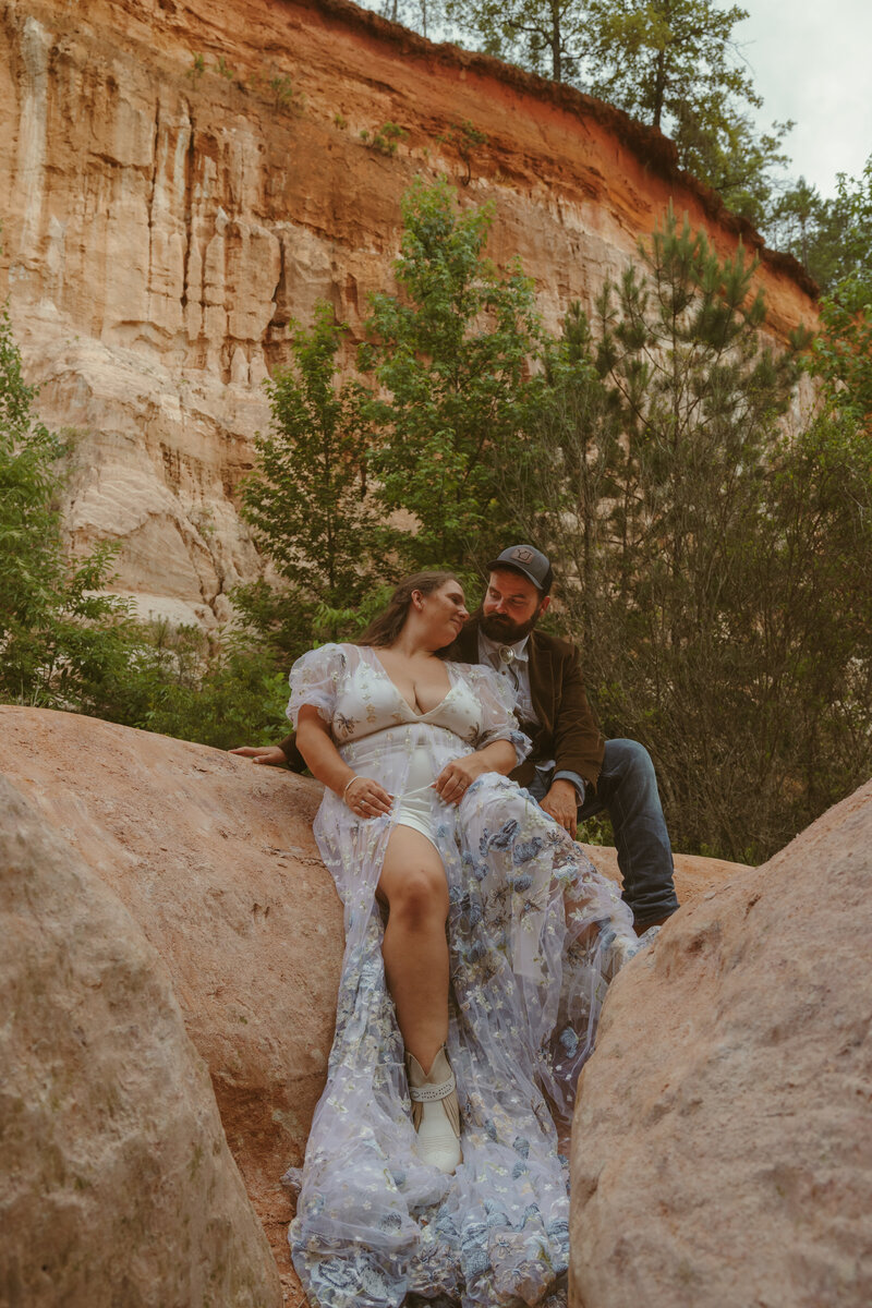 Couple sitting closely together on large canyon rocks surrounded by greenery, with the bride in a flowy floral gown and the groom in a jacket and cap, sharing an intimate moment during their adventurous canyon elopement.
