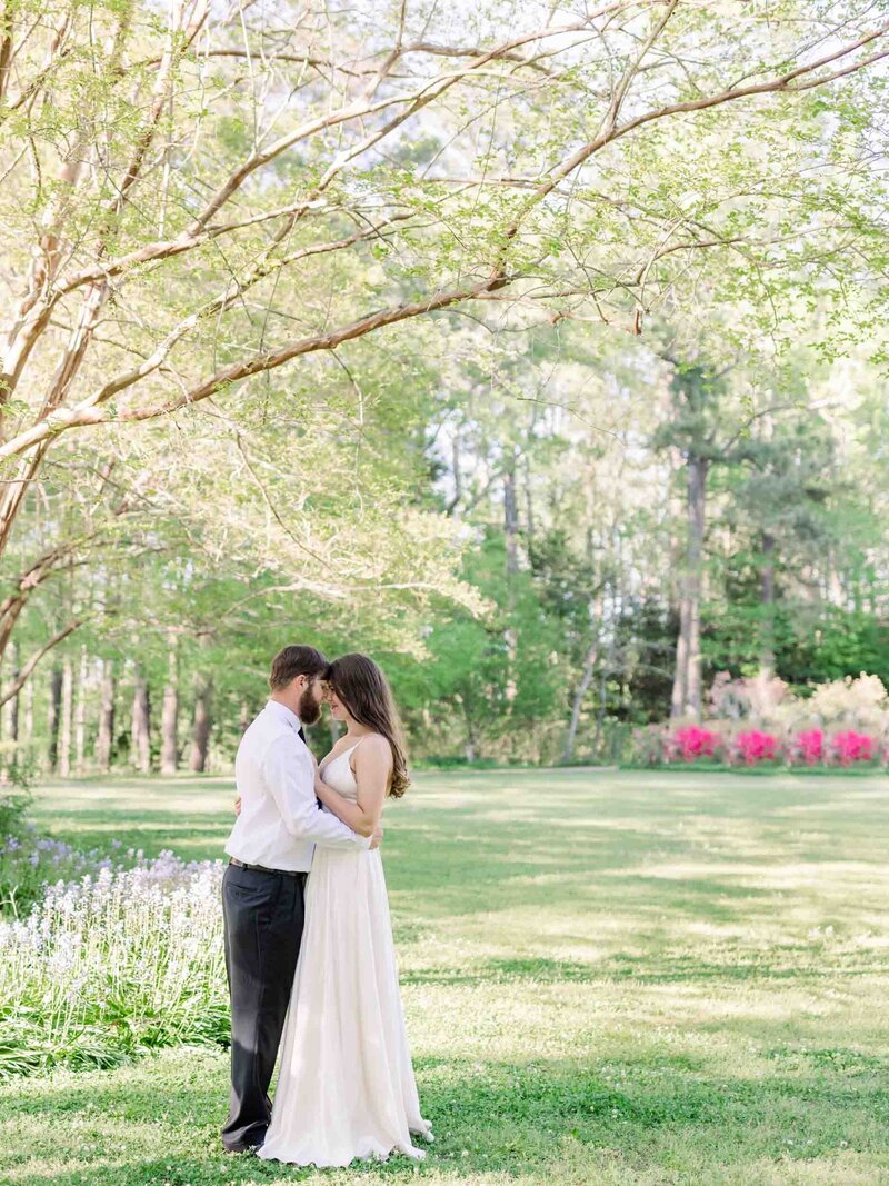 A man holding a woman in his arms under the canopy of a tree in a green field of grass and flowers during their scheduled portrait session  at Callaway Gardens.