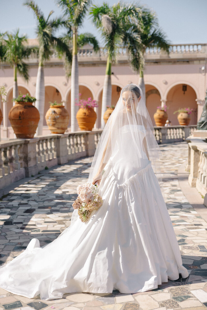 A bride wearing a voluminous white wedding gown and a sheer veil that covers her face. Colonnade of arches along stone railings. Ringling museum. 