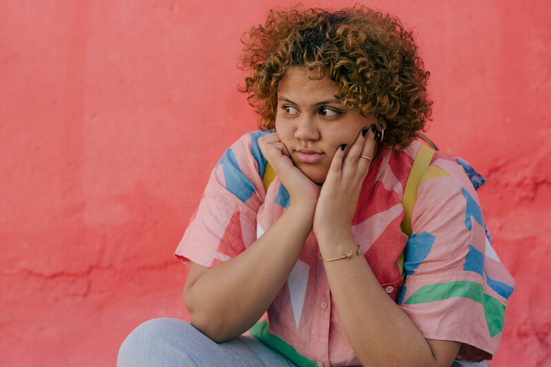 A teen with natural, curly hair sits against a brightly-colored wall, face in their hands. They look past the viewer, with a sad expression on their face.