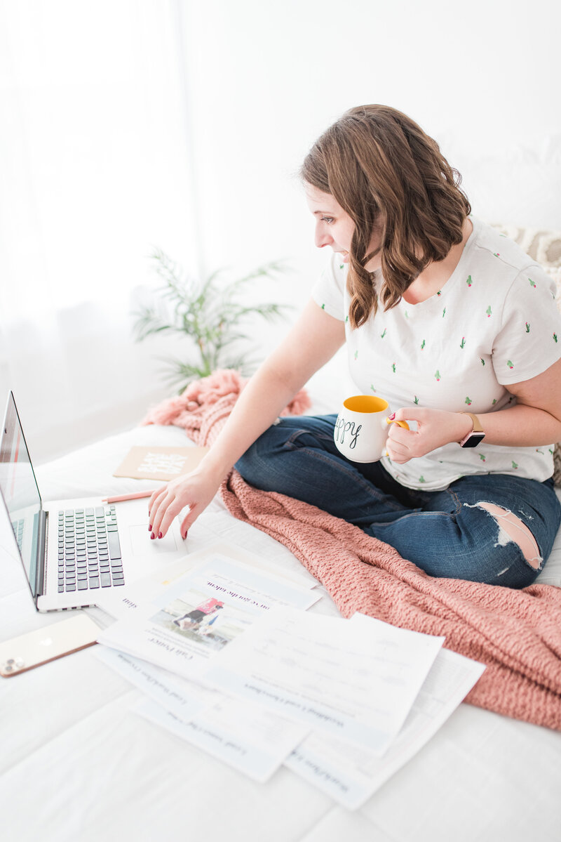 woman sitting on the bed with a mug on her hand and working on her laptop