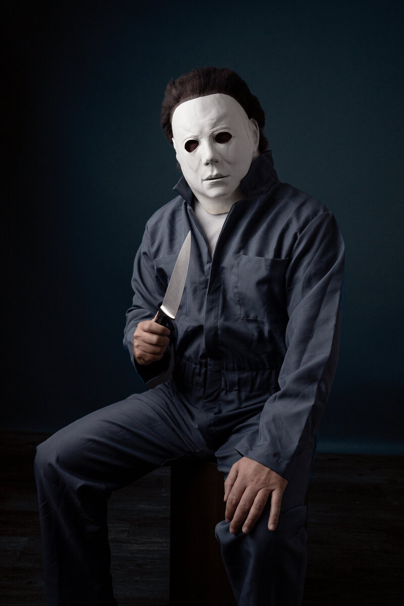 Man sitting on an apple box in a Michael Myers Costume holding a knife in his right hand. Image has blue color toning in Holly Springs, NC