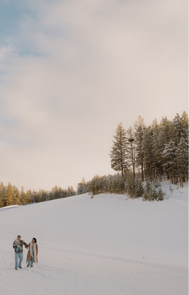 A husband and wife walk through the snow holding their baby in a family photo.