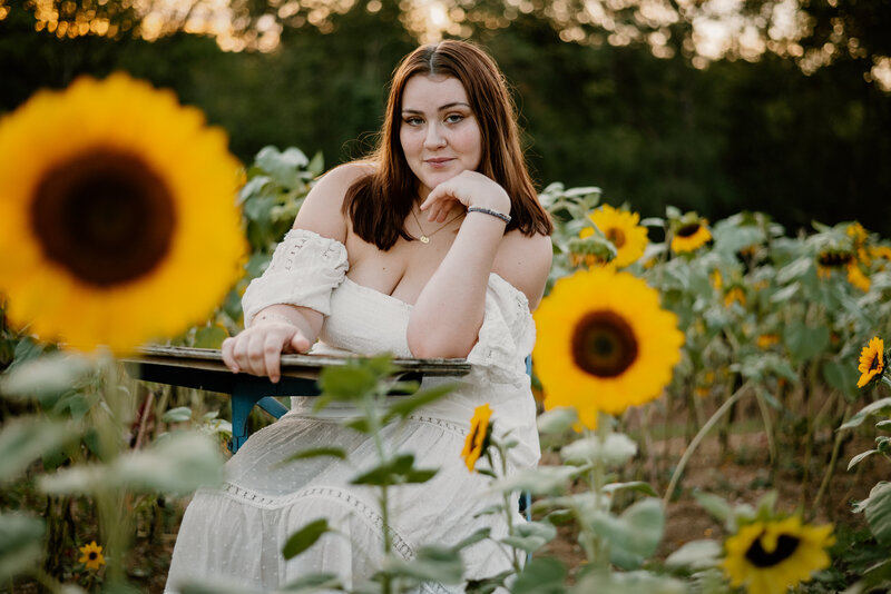 Senior photo of teen girl sitting at a vintage desk in a sunflower field smiling at the camera.