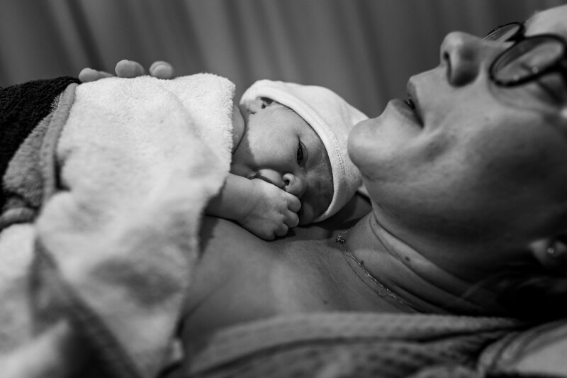 Moeder ligt bij te komen van de bevalling en haar dochter ligt op haar borst met een duim in haar mond.