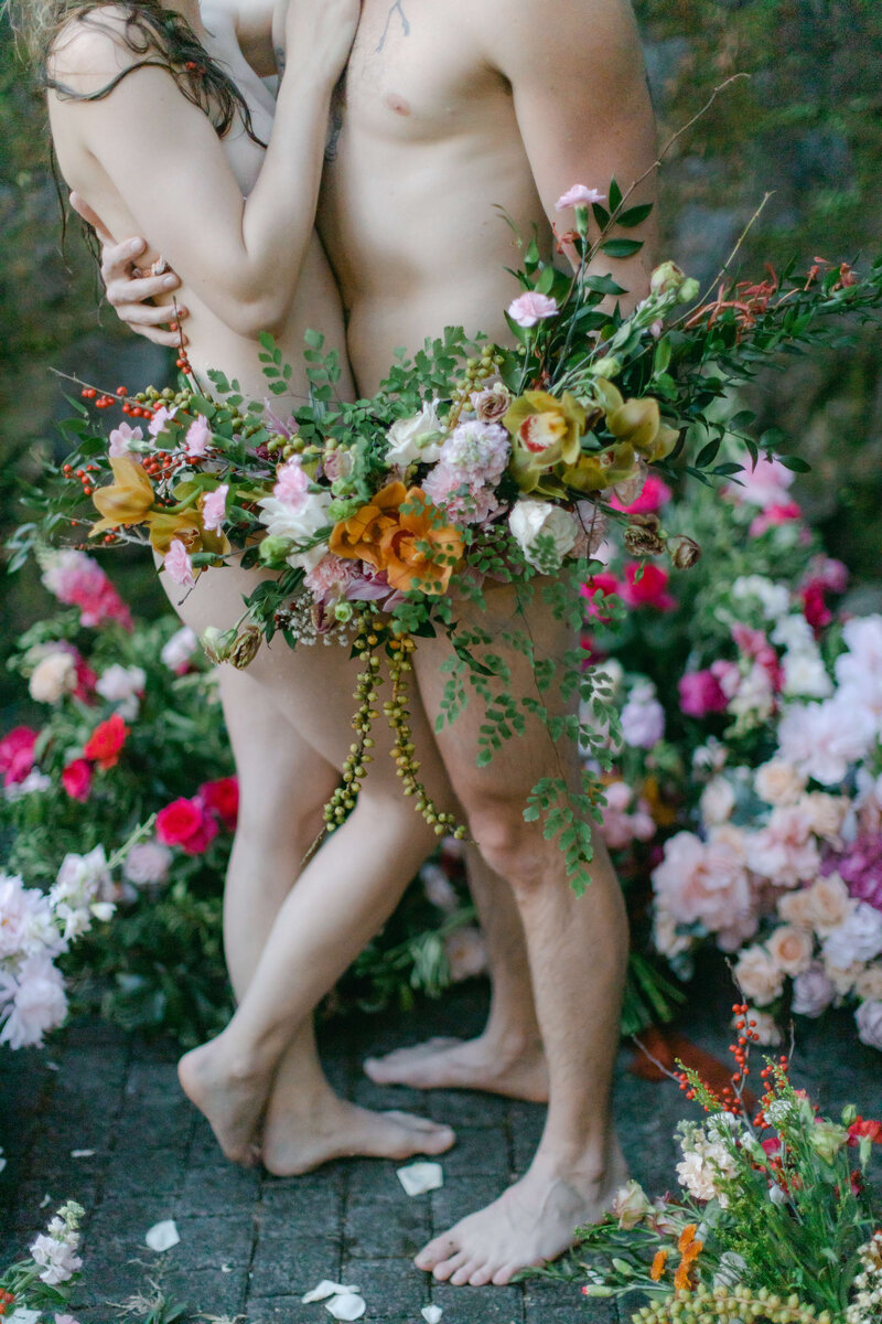 A bride and groom stand naked holding only flowers to cover themselves.