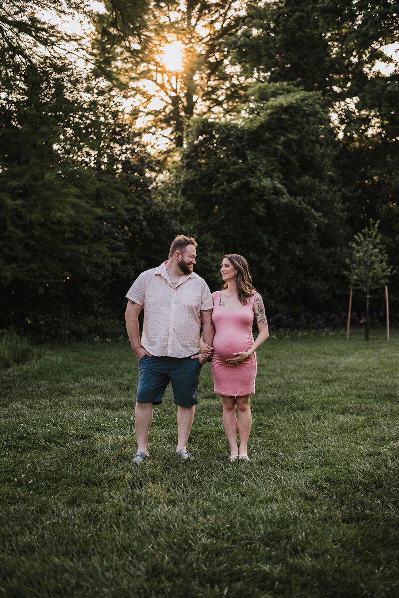 Dad in pink and blue standing in park next to pregnant mom holding her belly in pink dress looking at each other in Baltimore Maryland