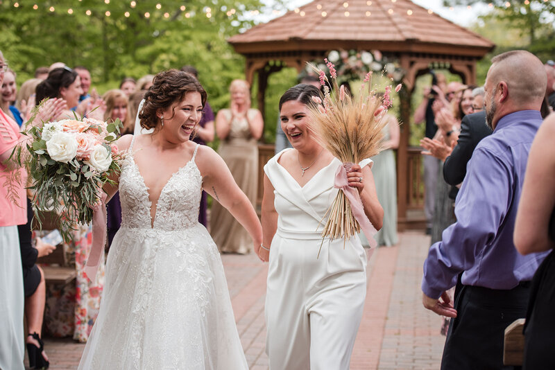 Two brides walking hand-in-hand, with one holding a bouquet and the other a bundle of wheat, as guests cheer at Pavilion on Crystal Lake