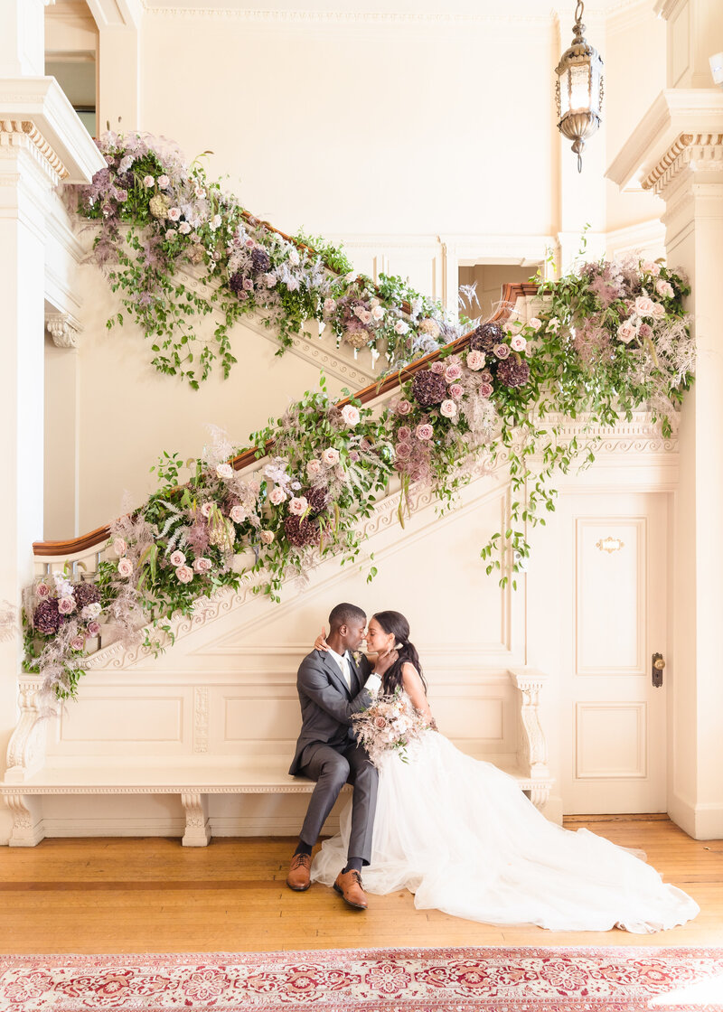 A bride and groom lean in for a kiss on a bench in front of a long staircase decorated with elaborate florals