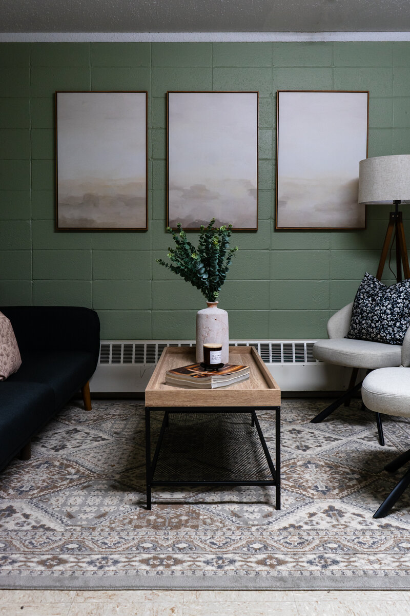 Commercial office space located inside a senior assisted living facility with sage green walls, calming art pieces and a dark sofa, coffee table and two additional chairs for a comforting space for families to sit with director.