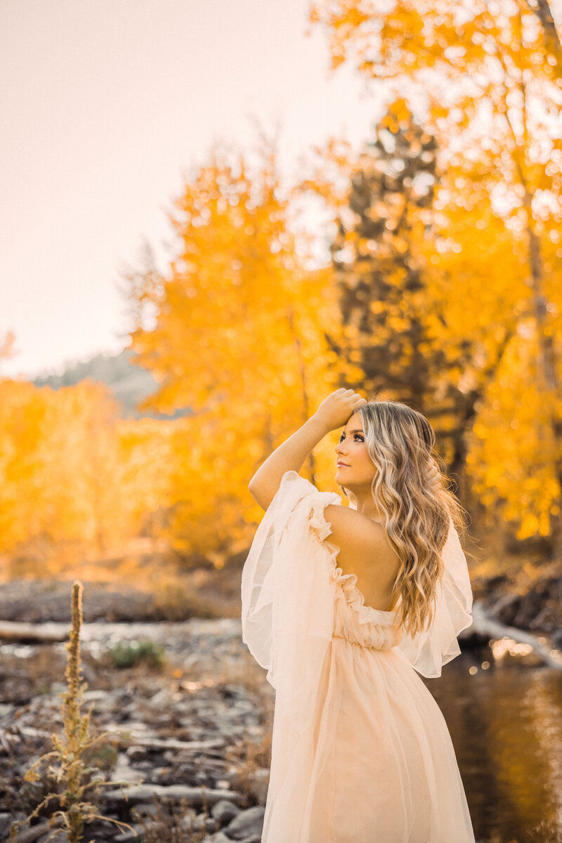 senior girl with long light hair standing by a river with yellow trees