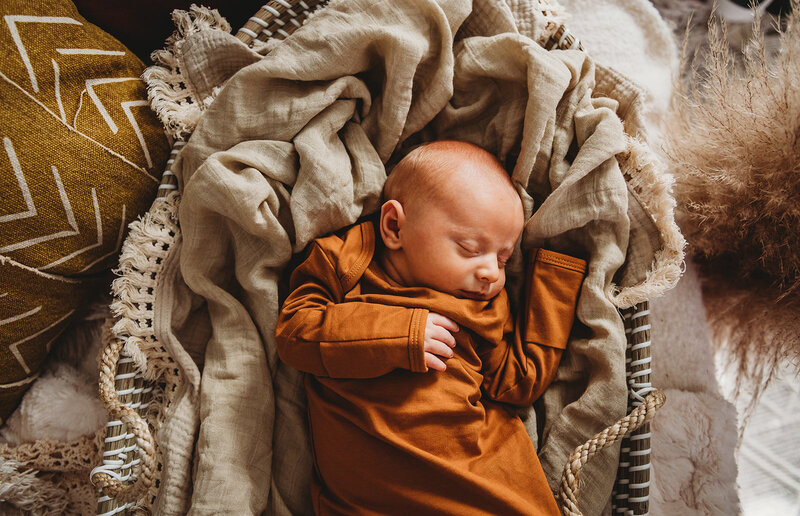 Family photographers Maryland photographs newborn photos with little baby boy in a burnt orange onsie laying in a basket with a beige blanket