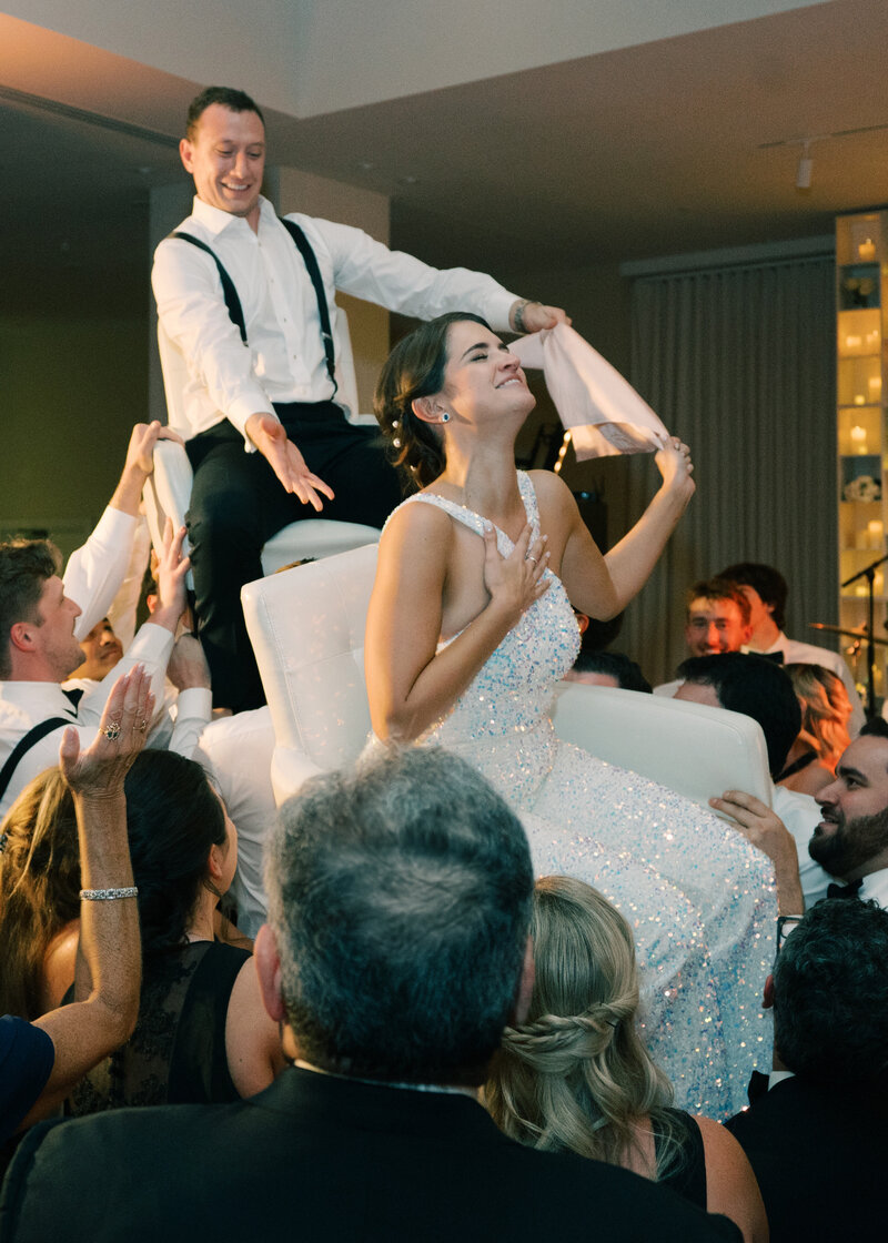 bride and groom in chairs lifted in the air during the hora dance at their wedding in DC photographed by wedding photographer Omar & Co