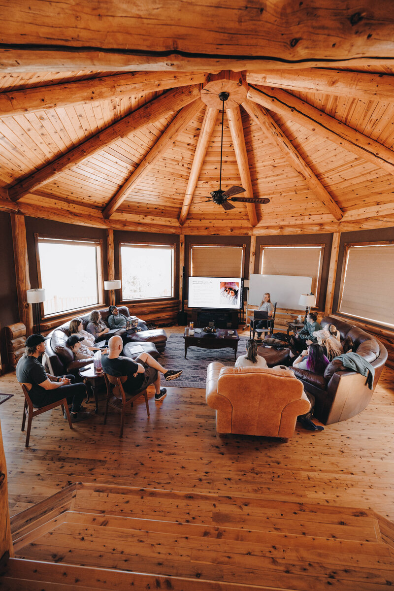 A group of people sit around a living room in a beautiful cabin in the woods.