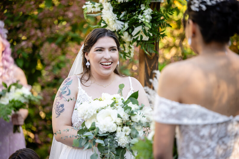 curvy white bride in white spaghetti-strap gown holding a white bouquet at the ceremony altar smiling at her bride