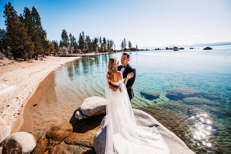 A newlywed couple stands on smooth boulders along the shoreline of Lake Tahoe, gazing at each other with the crystal-clear turquoise water and a sandy beach stretching behind them.