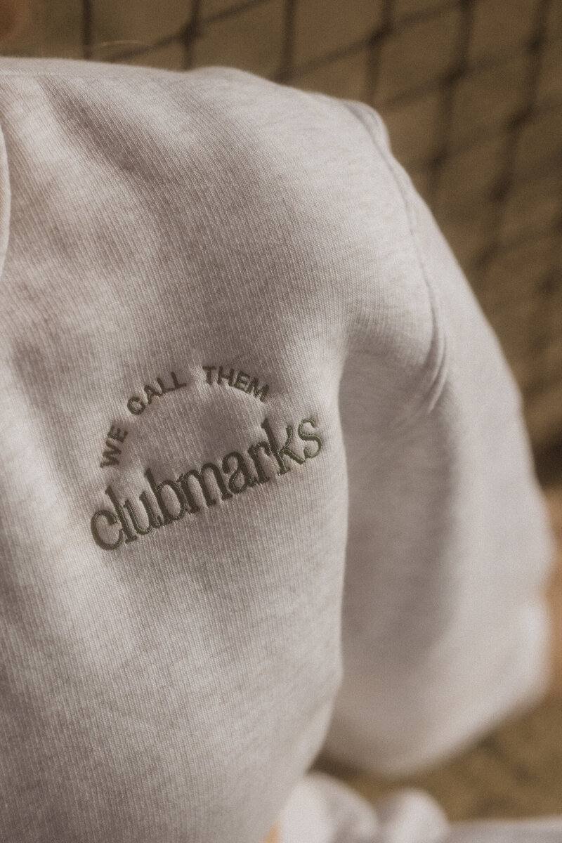 Close-up of embroidered “We Call Them Clubmarks” logo on soft beige fabric, highlighting brand identity and apparel design.