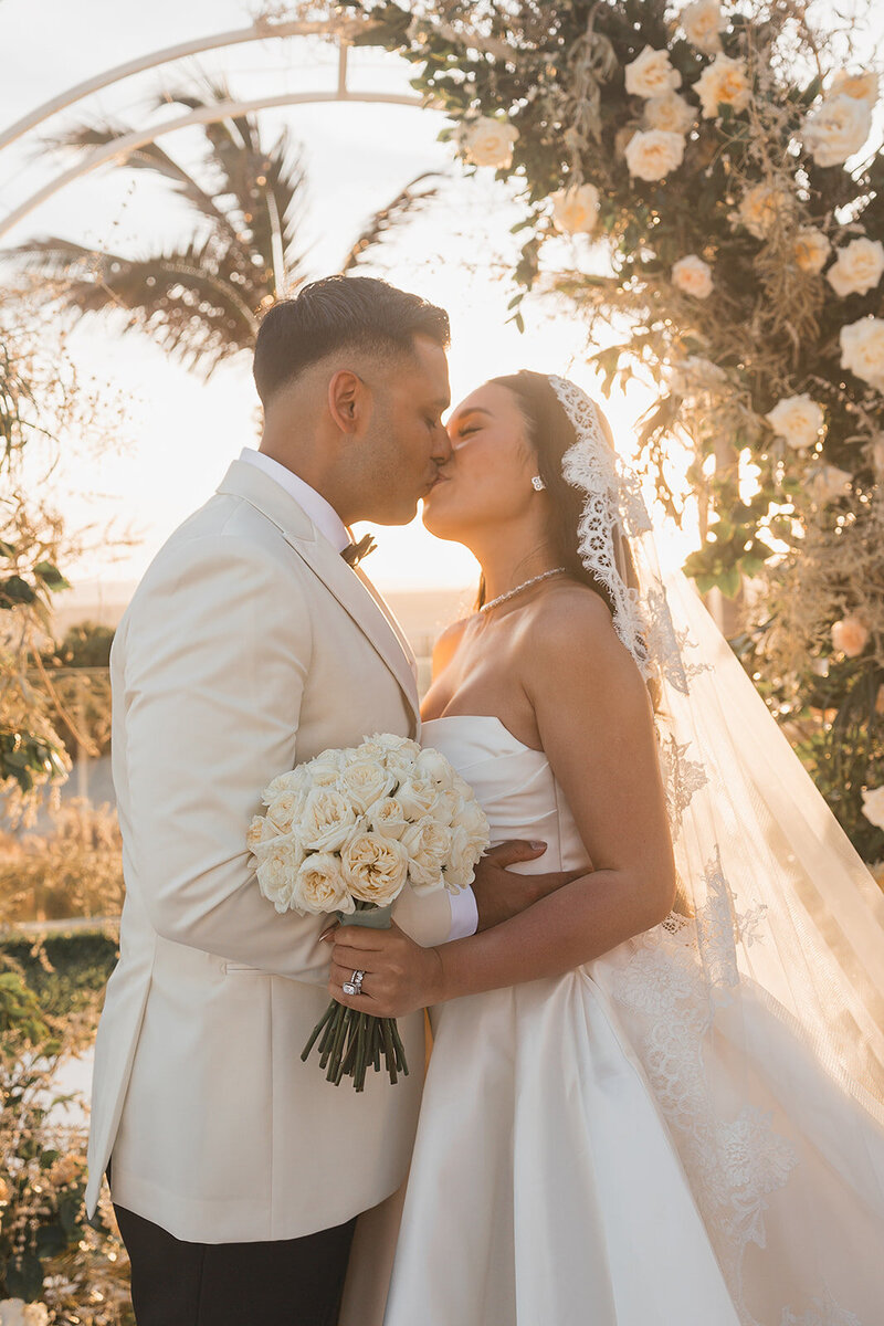 Bride and groom kiss while holding bouquet of white flowers in front of sunset
