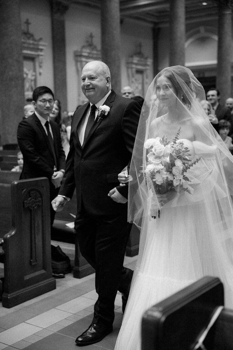 Father walks his daughter down the aisle of a cathedral during her wedding ceremony. 
