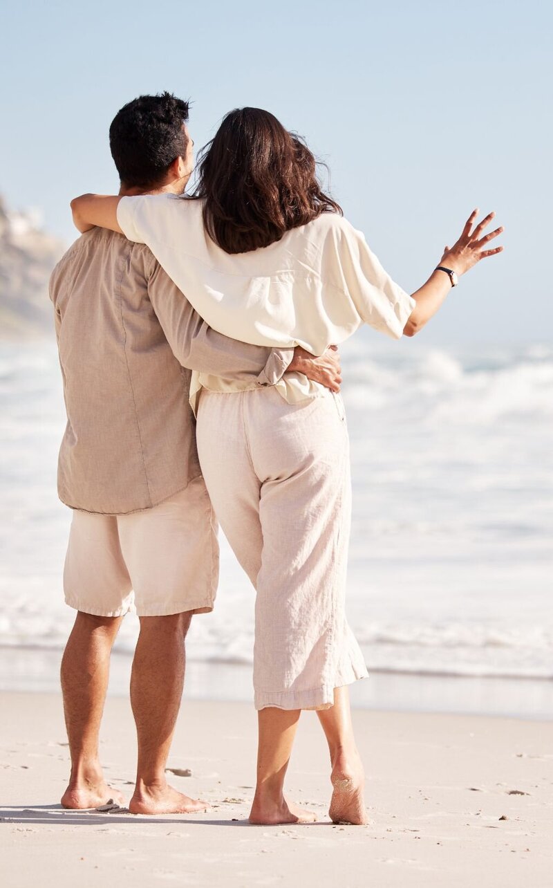 A loving couple hugging on the beach and looking at the waves represents how married couples can heal from infidelity with The Affair Recovery Online Program: It's Okay To Stay by Relationship Experts