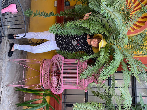 Author Beth Harkins in a yellow sun hat under a fern next to a pink wire cage.