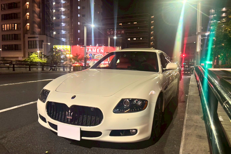Front view of a white Maserati Quattroporte parked on a city street at night in Tokyo, illuminated by urban lights and surrounded by modern buildings, showcasing its sleek design and signature grille.