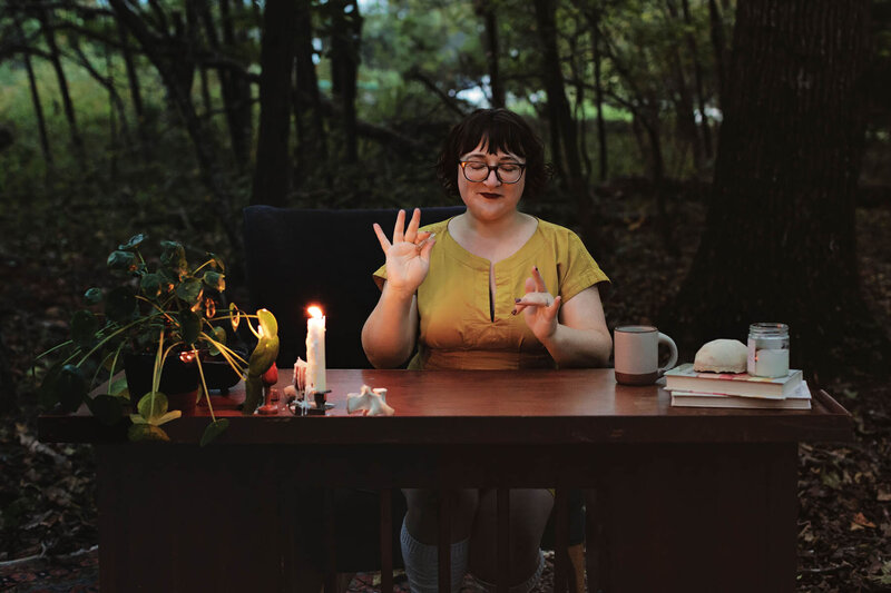 Ash Stinson sits at a table with a plant, lit candle, and stack of books while practicing energy work in the forest
