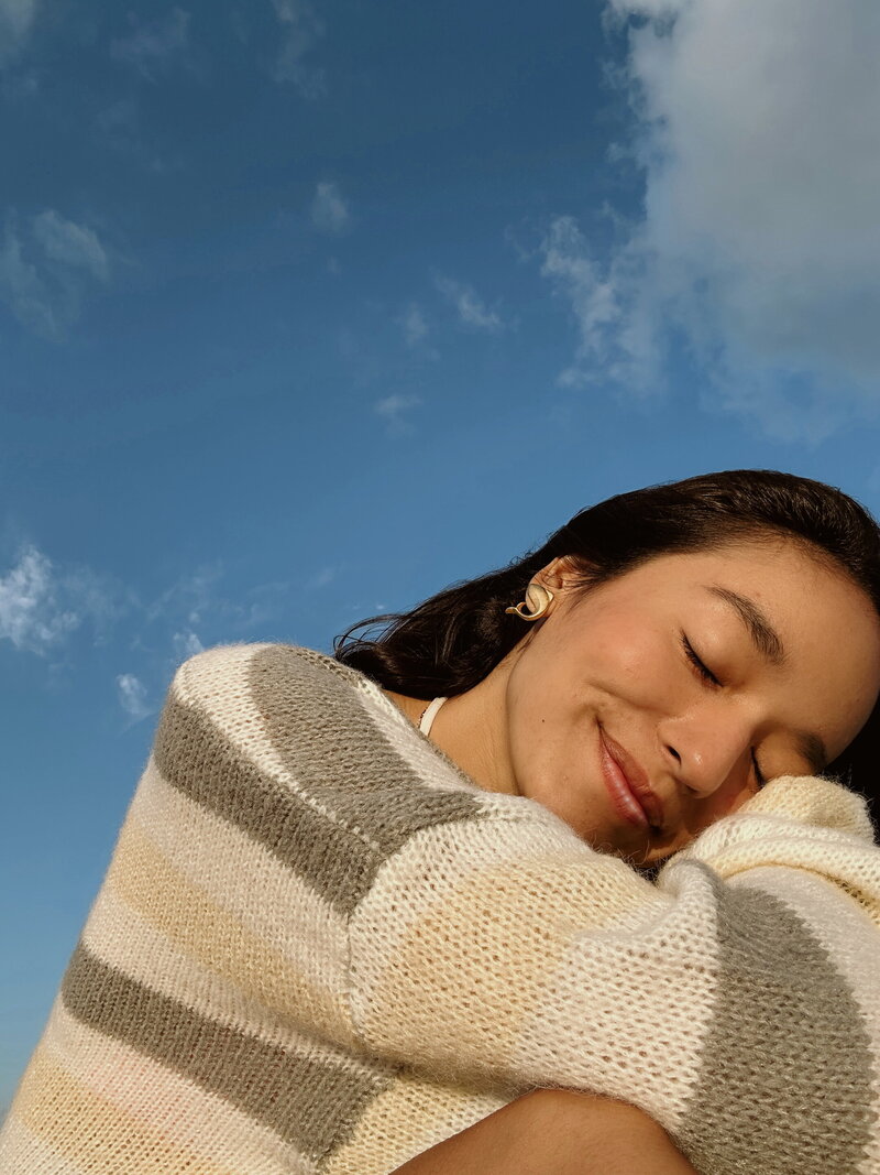 A woman with closed eyes and a peaceful smile, wearing a striped sweater against a blue sky with few clouds. The mood is serene and content.
