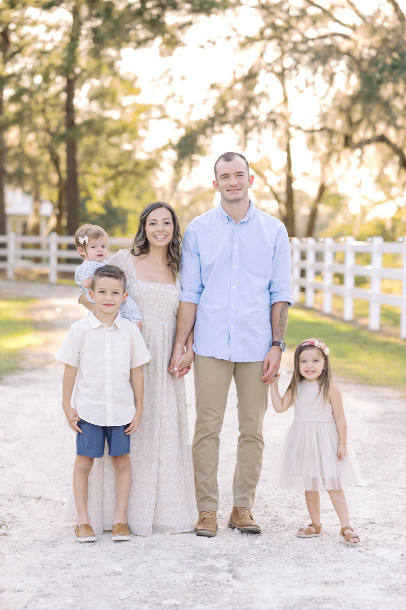 Lifestyle family session on a dirt path with sunlit trees and white fencing.