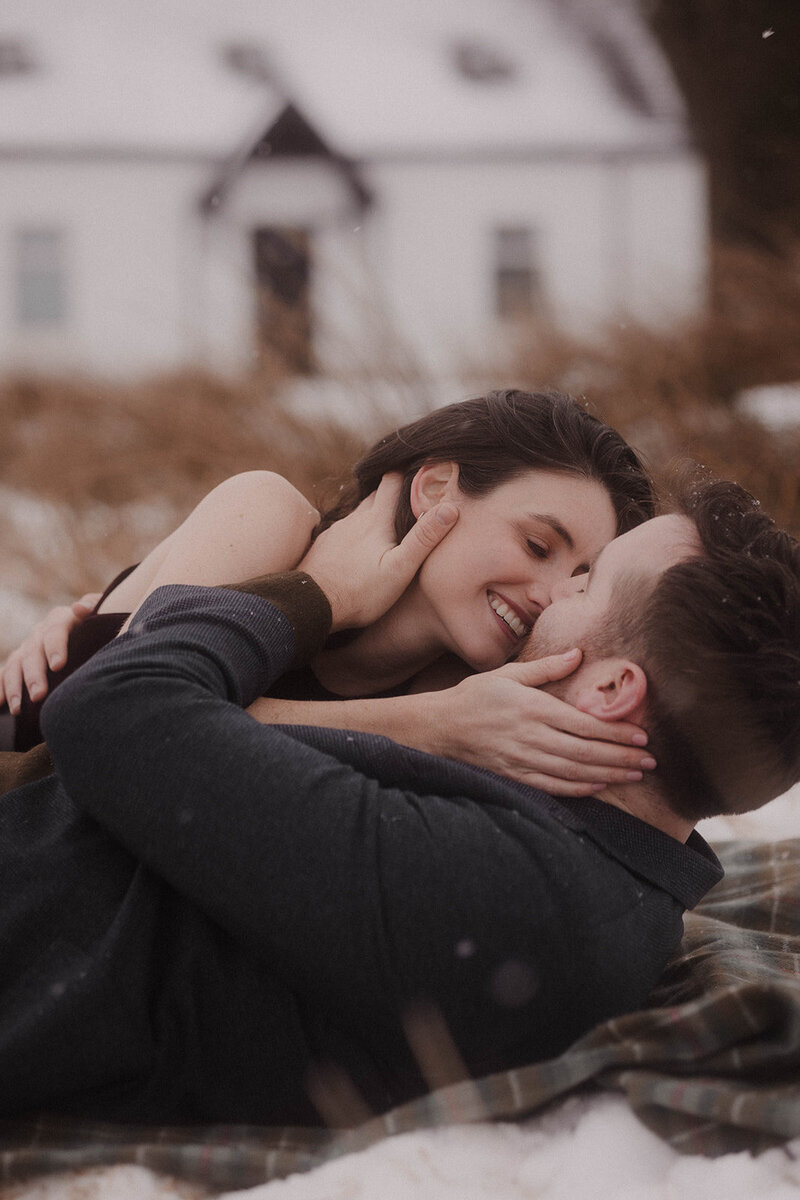 Romantic winter couples session in the Scottish Highlands, with a pair laughing and embracing in the snow outside a white cottage, captured in a cinematic fine art style by wedding photographer Aly Robinson.