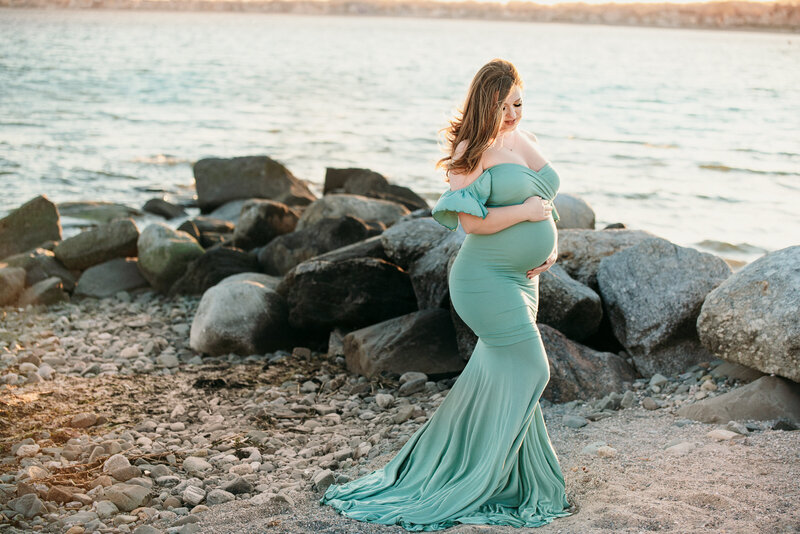 pregnant woman in seafoam green dress posing on beach looking down holding belly