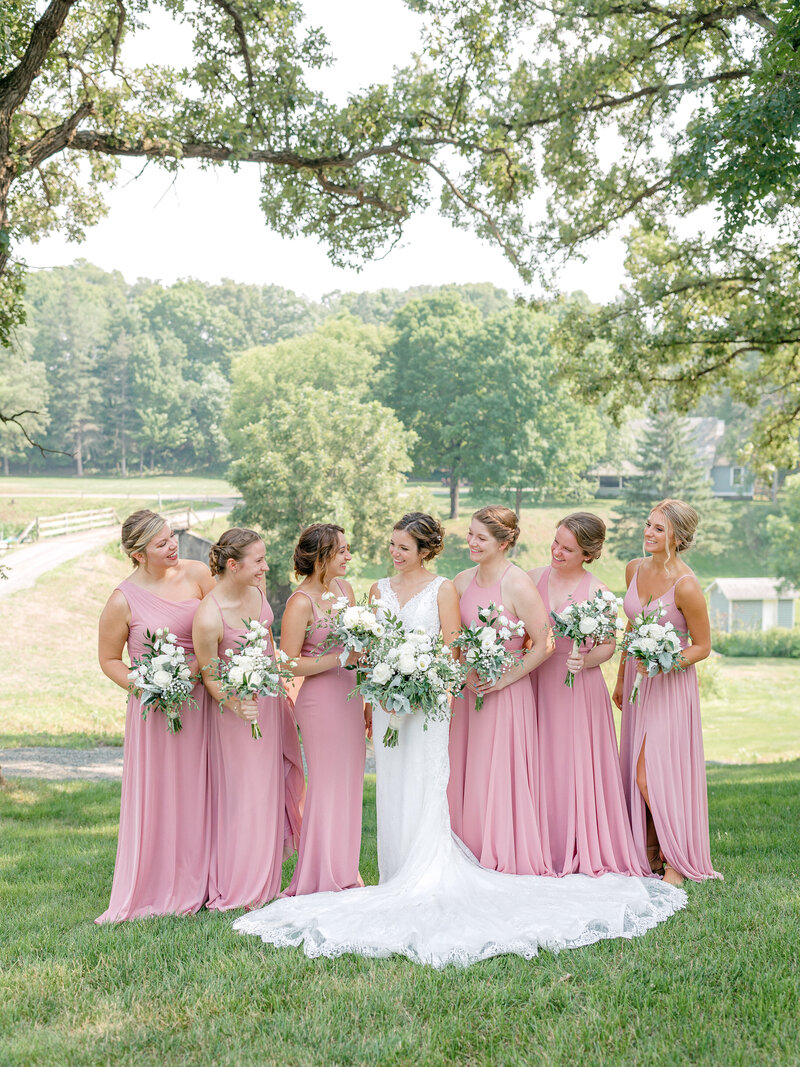 Bride and her bridesmaids in pink dresses standing outside under trees and smiling at each other