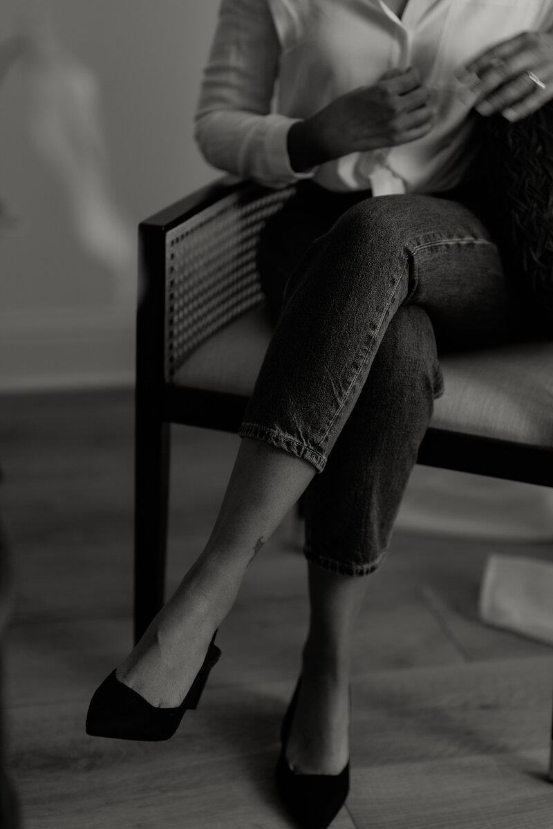 Black and white branding photo featuring a close-up of a Black woman photographer's fashionable suede pointy slingback shoes, seated in a chair in her Fort Worth photo studio. The image highlights her stylish and professional demeanor