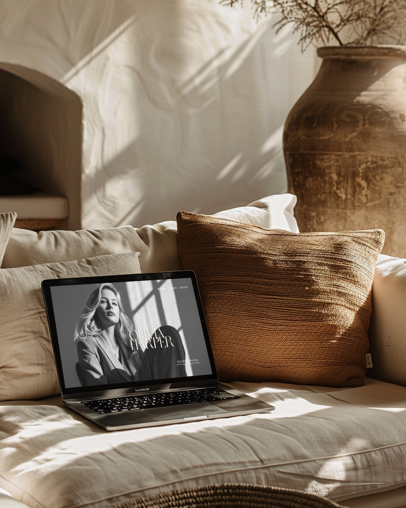 Laptop mockup featuring a black and white brand visual, styled on a sun-drenched beige couch with textured throw pillows and earthy ceramic décor—capturing warm, modern CEO vibes.