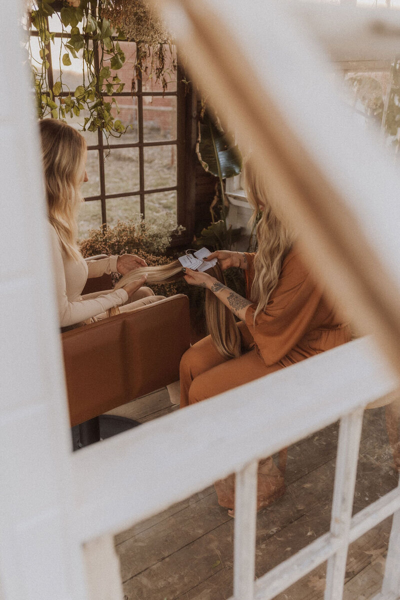 The hair extension specialist in Dallas, Texas is showing the product to the client who is sitting in a brown salon chair. Behind them is a wooden window with a view of greenery and they are dressed in brown and orange outfits.