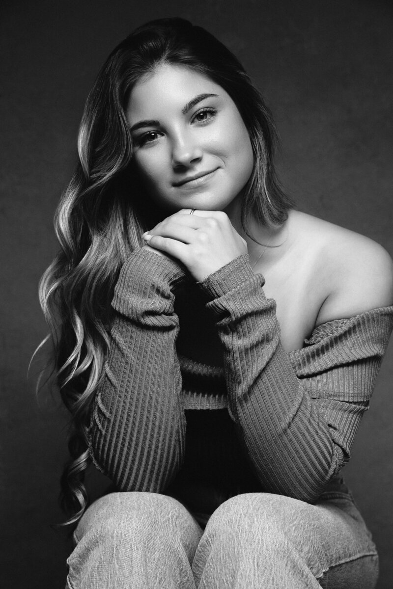 Black and white senior portrait of girl sitting with overlapping hands under her chin and her elbows up on her knees. She is smiling and has wavy hair cascading down her right side.