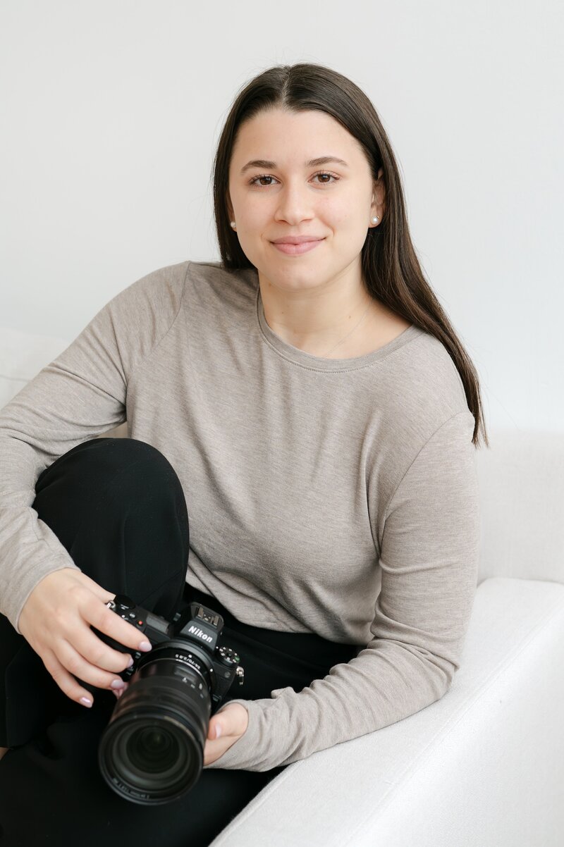 Ephe sits on a white couch with her leg near her chest and holds her Nikon camera.