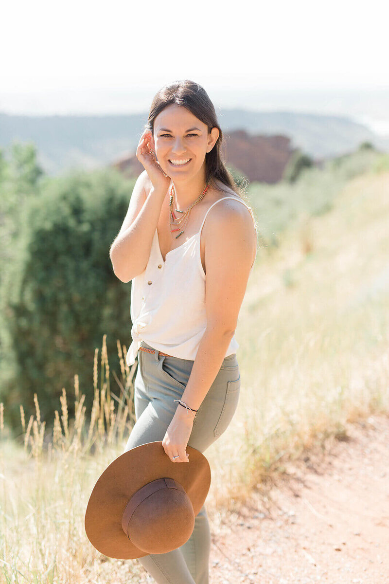 Laura, Founder of Find Your Freedom Co., standing in the sunchine smiling at the camera and holding a hat.  She is wearing a white tank top, green pants, and pink, gold, and green beaded necklaces.